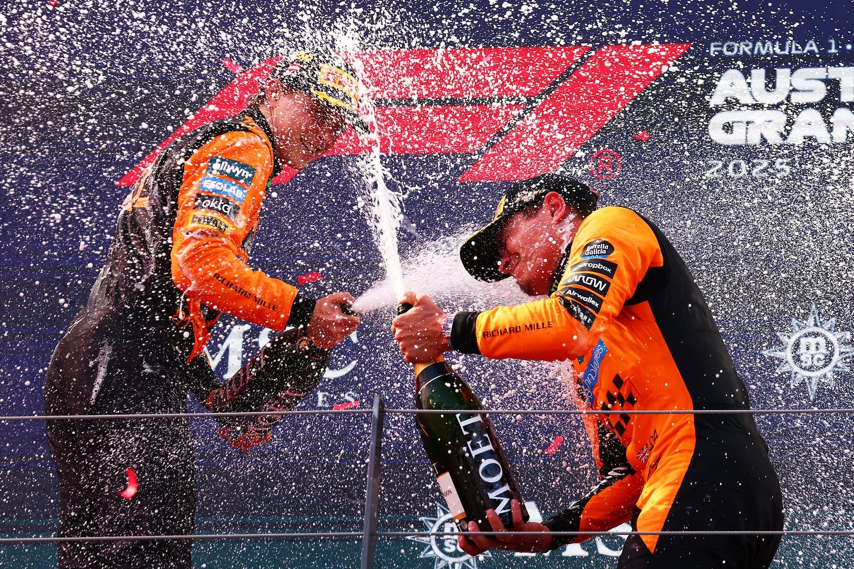 Two race car drivers wearing orange and black racing suits celebrate on a podium by spraying champagne, with bubbles and liquid flying in the air. The background shows event signage for the Formula 1 Australian Grand Prix 2023.
