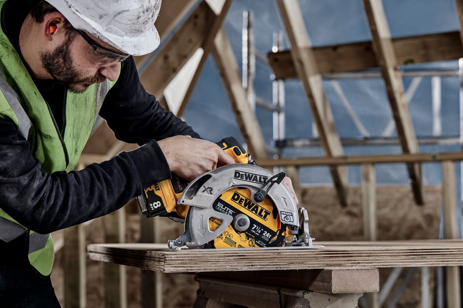 A person wearing a safety helmet and vest is using a DEWALT circular saw to cut wooden boards at a construction site with exposed wooden beams in the background.