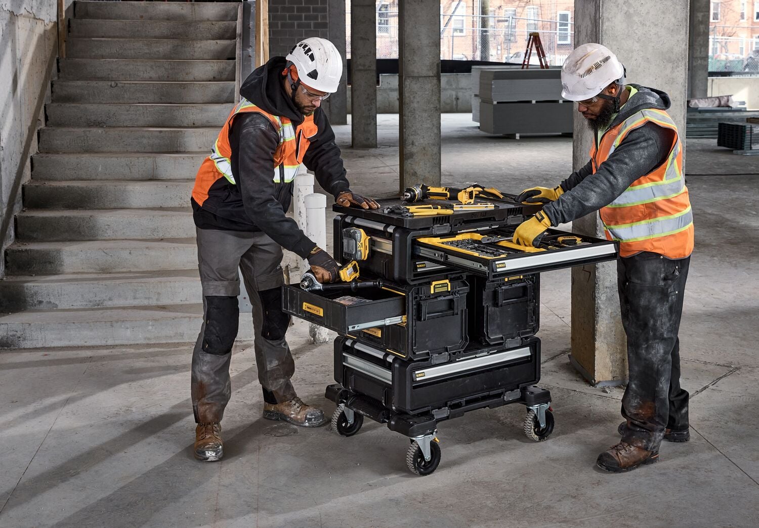Two construction workers wearing safety vests and helmets are using a DEWALT rolling tool storage cart with multiple drawers and compartments in a partially finished building. Various DEWALT tools are placed on the cart.