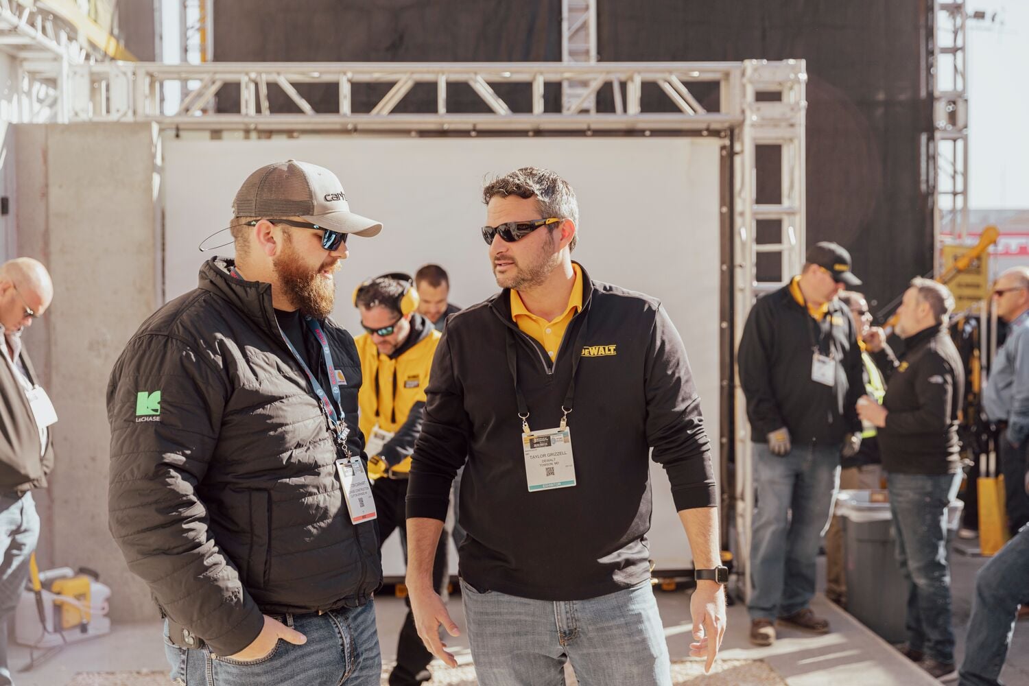 A group of people wearing branded jackets and name tags are gathered at an outdoor event or trade show. The setting includes a metal framework structure and several people appear to be engaged in conversation.