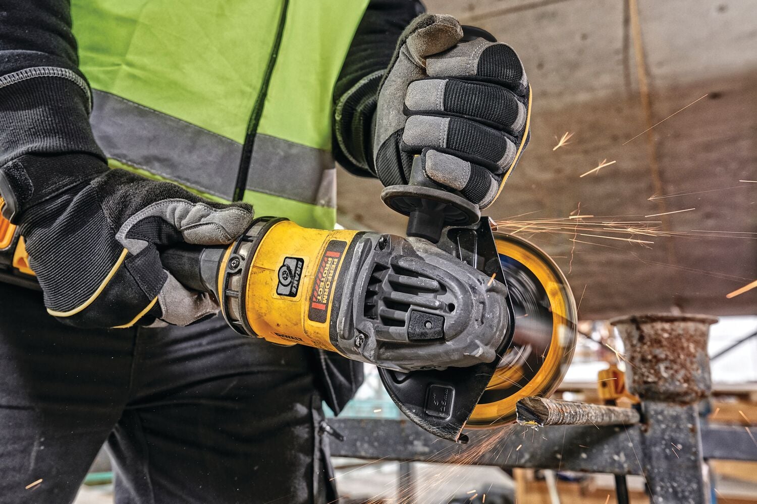 Close-up of a person wearing safety gloves and a reflective vest, using a yellow angle grinder to cut a metal pipe. Sparks are visibly flying from the cutting area.