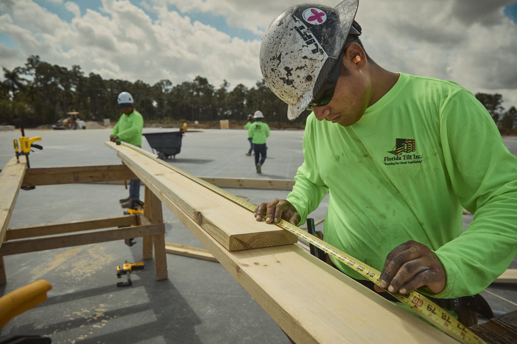A construction worker wearing a neon green shirt and a hard hat measures a long wooden plank using a tape measure at an outdoor construction site. Two other workers in similar attire are seen in the background, and various construction tools are visible on the wooden structure.