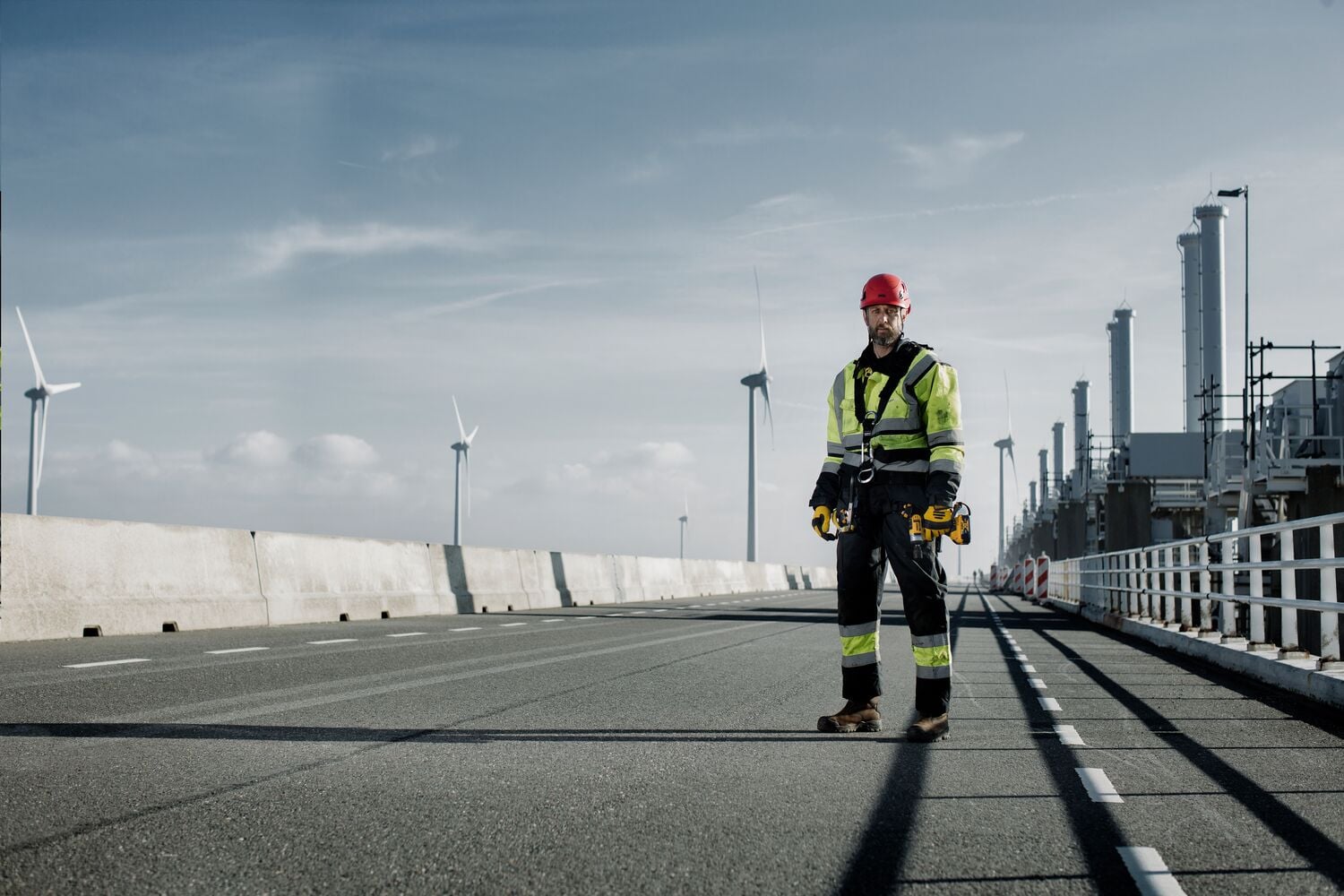 A worker wearing high-visibility protective clothing and a red helmet stands on an industrial road, holding DEWALT tools. Wind turbines and factory structures are visible in the background on a clear day.