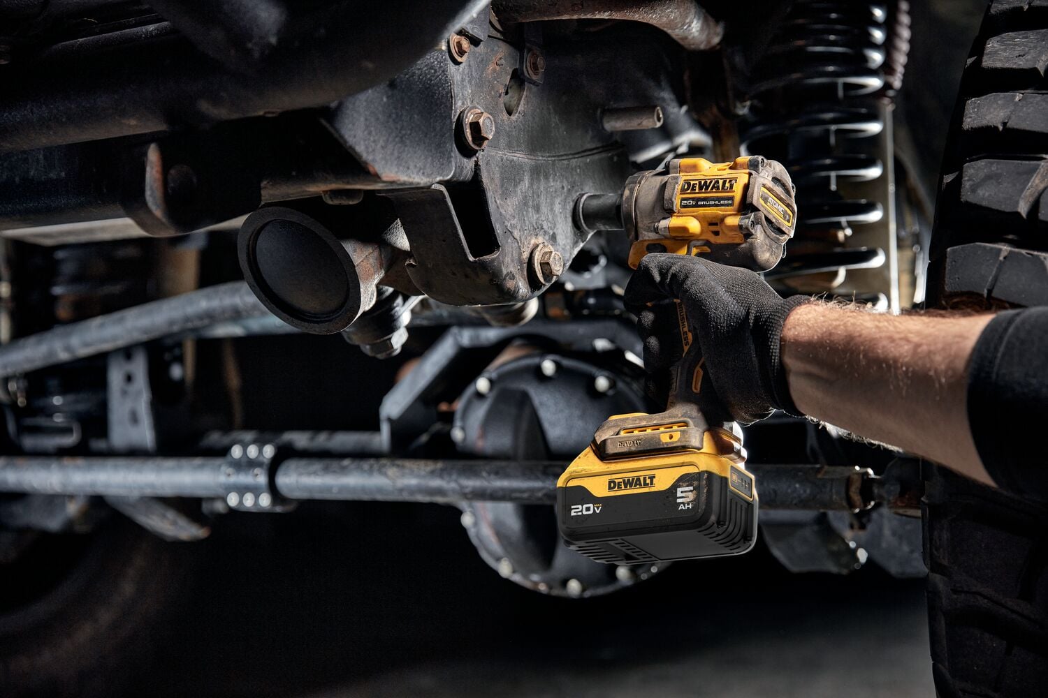 A person wearing a black glove uses a DEWALT 20V cordless impact wrench to work on the undercarriage of a vehicle, specifically tightening or loosening bolts on a metal component.