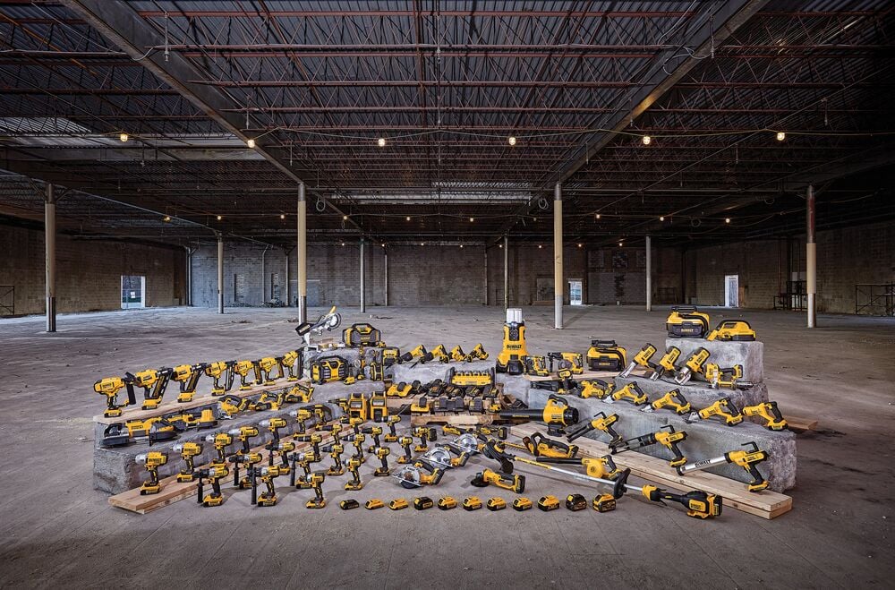 A wide selection of yellow and black DEWALT power tools, batteries, and accessories are displayed on concrete blocks and wooden planks, arranged inside a large, empty warehouse with exposed ceilings and pillars.
