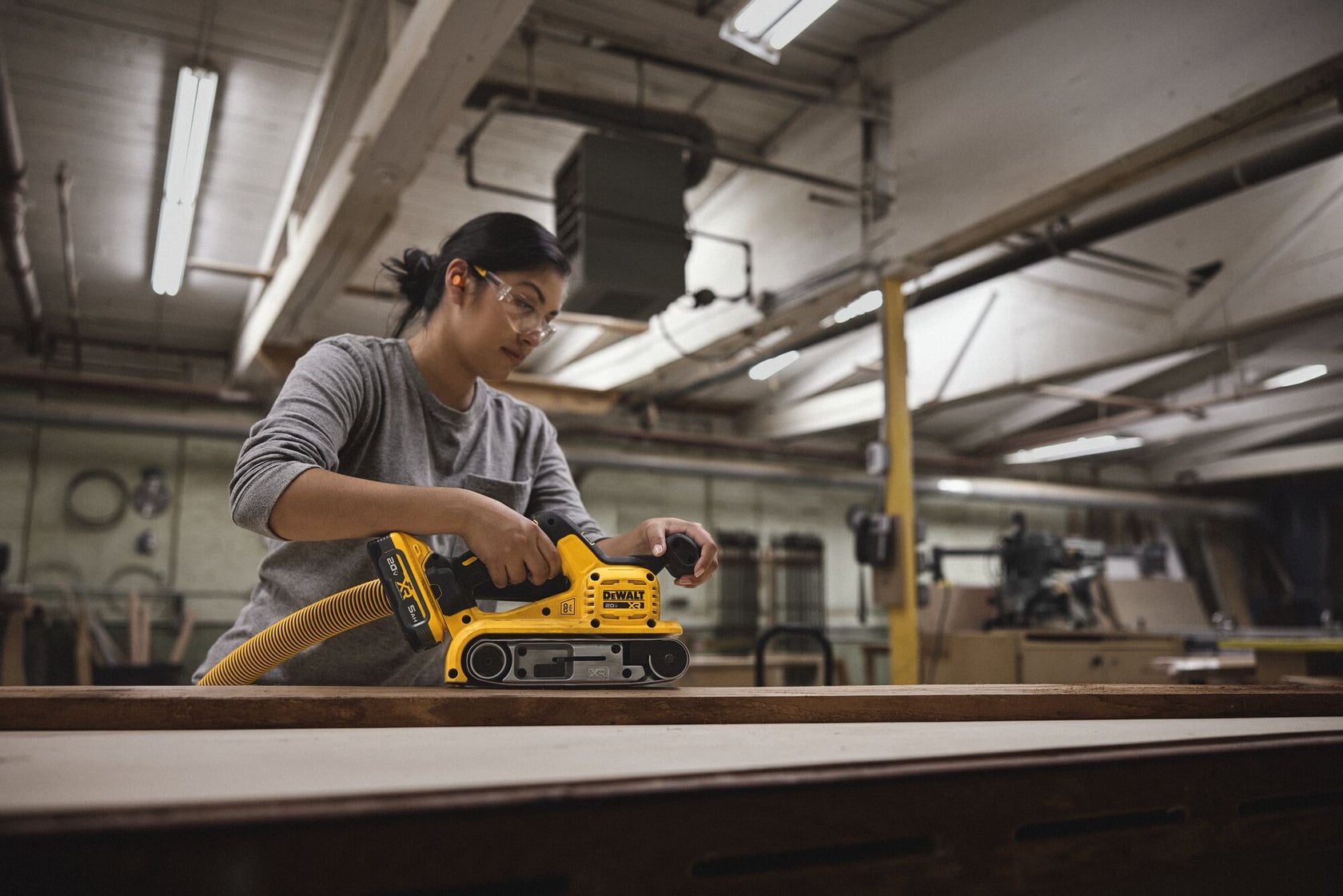 A person uses a DEWALT DCW220B belt sander on a wooden surface inside a workshop. The individual's face is blurred, and the workspace is filled with various tools and machinery.