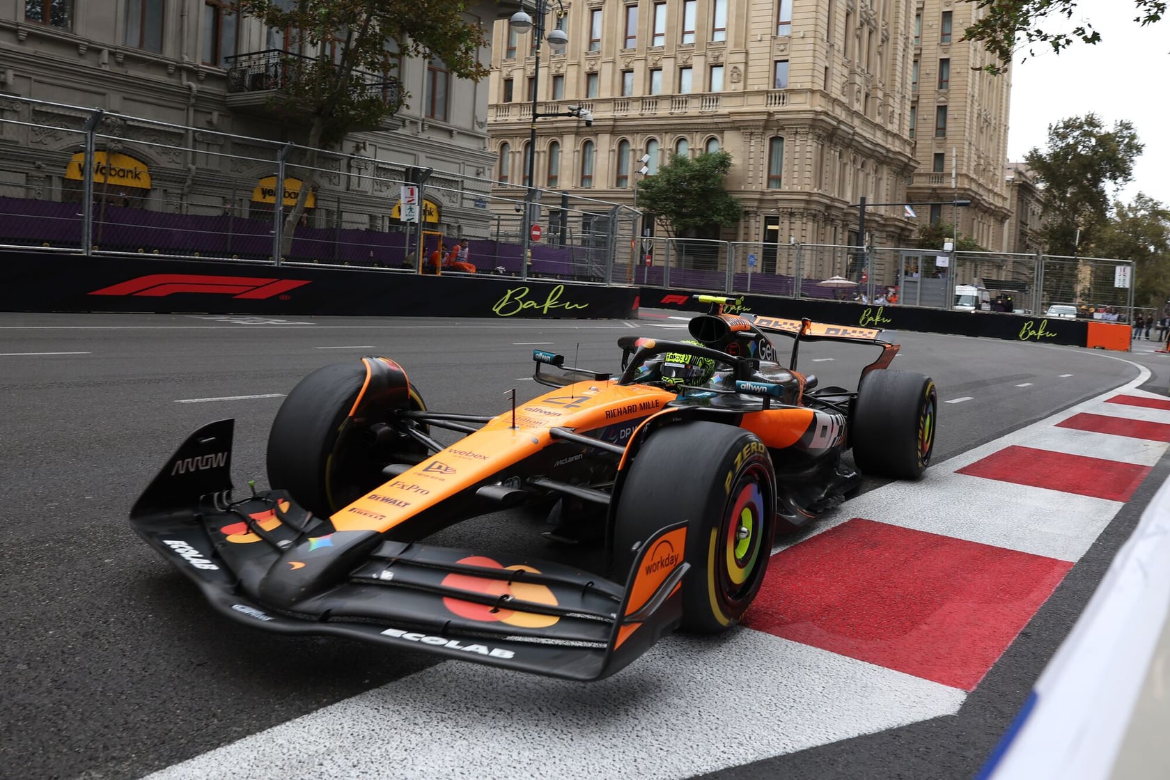 An orange and black Formula 1 race car driving on a street circuit during a race event in Baku. The city buildings and safety barriers are visible in the background.