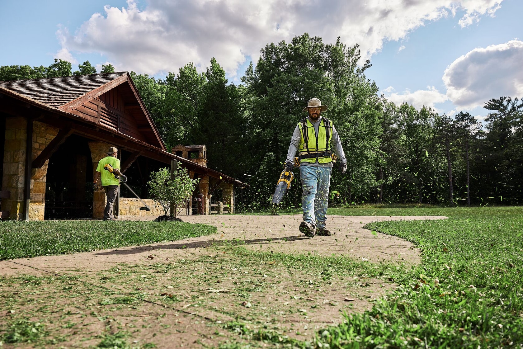 Two workers use DEWALT outdoor tools to clear grass near a house, with trees and cloudy sky in the background.