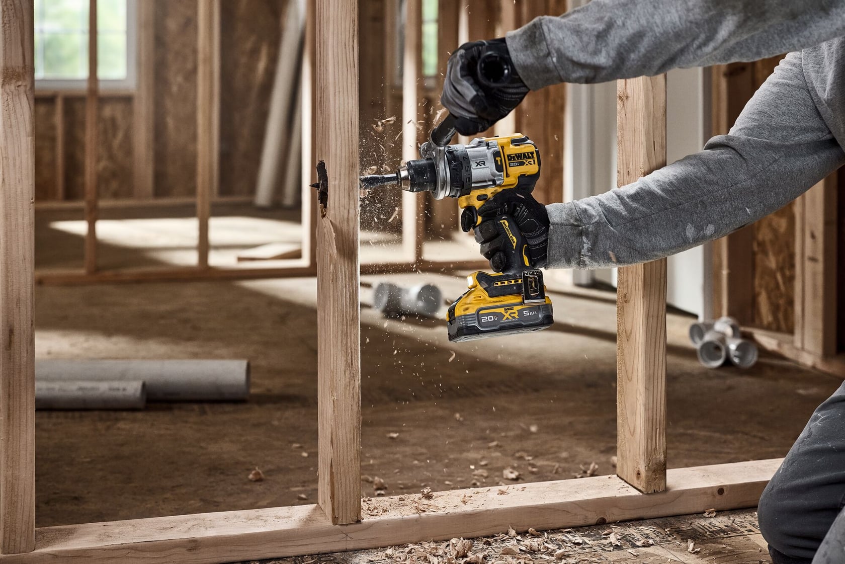 A person wearing gloves uses a DEWALT cordless drill to bore a hole in a wooden framing stud inside a building under construction. Wood chips are visible as the drill operates.