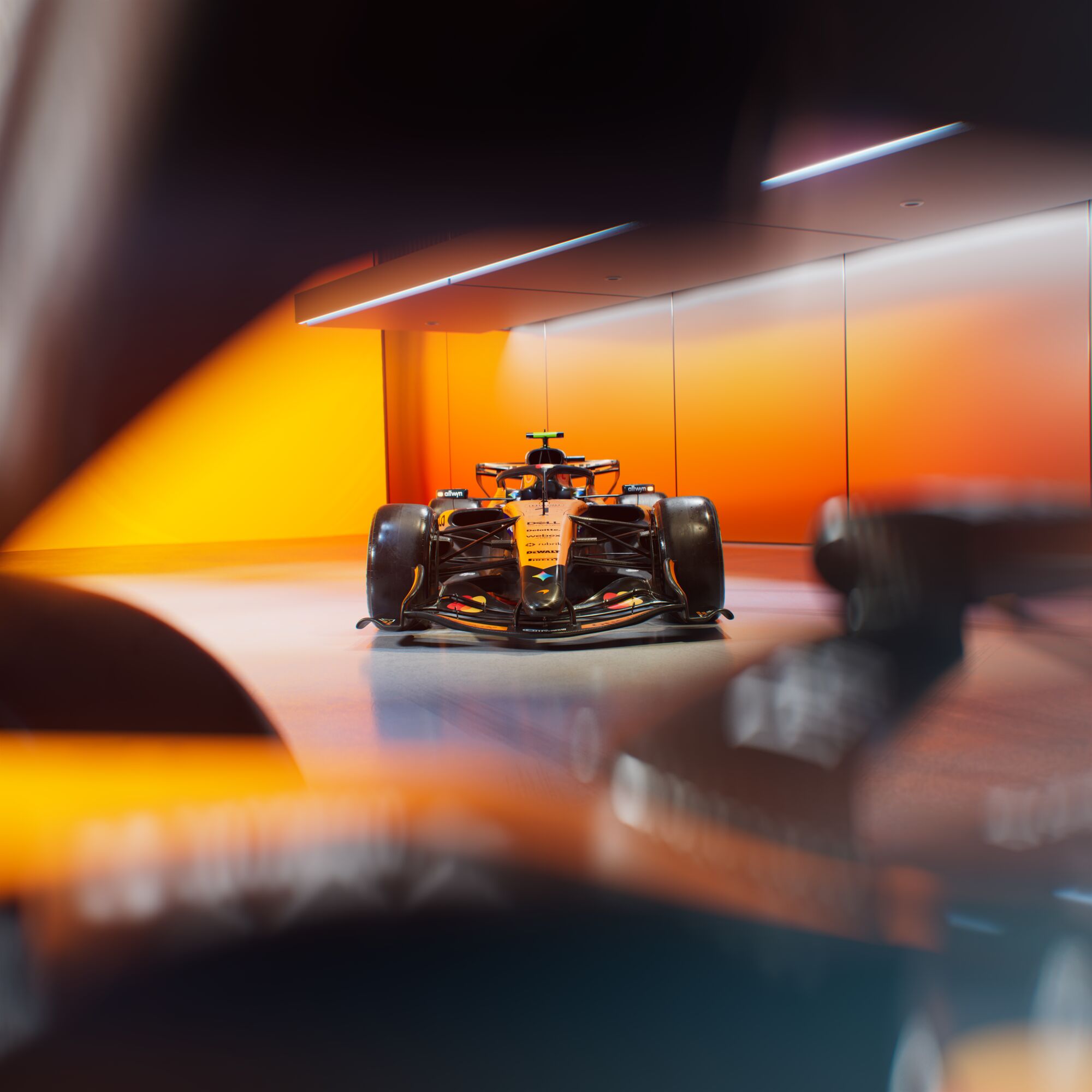 A modern Formula 1 race car with an orange and black color scheme is displayed in a sleek, brightly-lit room with orange walls. Part of another car is visible out of focus in the foreground.
