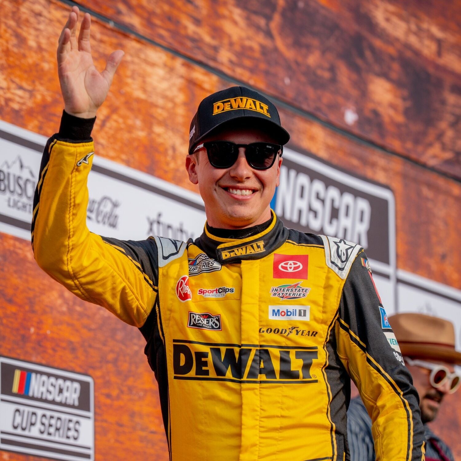 A person wearing a yellow DEWALT racing suit and cap is waving while standing in front of a NASCAR Cup Series event backdrop. Visible sponsor logos are displayed on the suit, and the background includes signage indicating the location as Nashville.