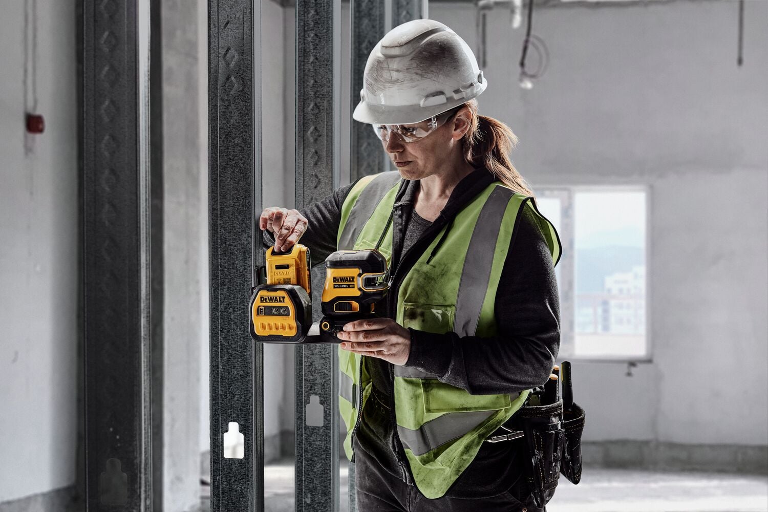 A construction worker wearing a hard hat and reflective vest operates a DEWALT laser level tool at a building site. The tool is yellow and black, and the worker is in an unfinished room with metal studs and construction materials.