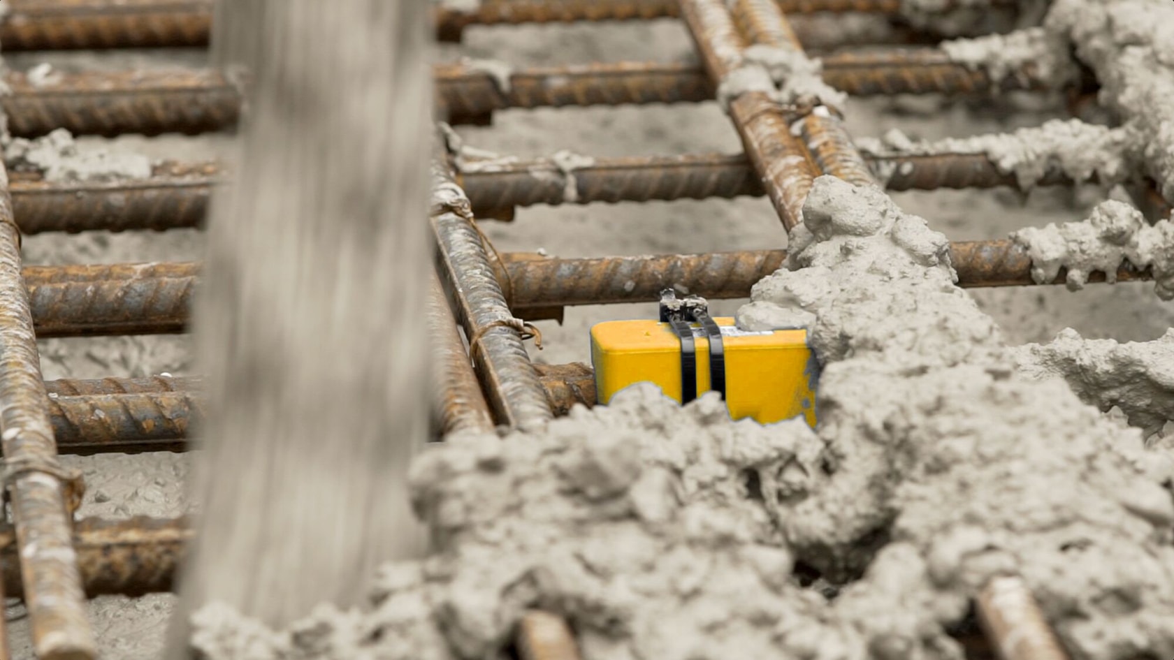 A yellow DEWALT device partially covered in wet concrete, positioned between metal rebar grids at a construction site.