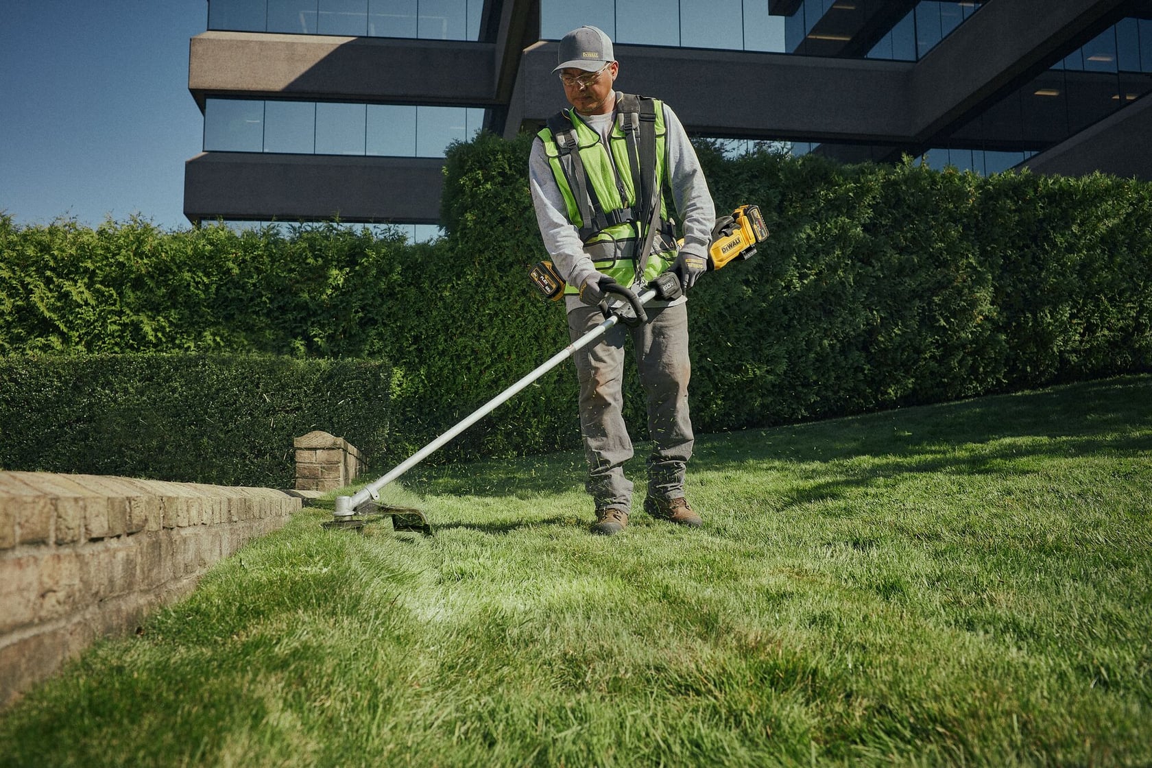 A person wearing a safety vest and gloves is using a DEWALT cordless string trimmer to edge the grass along a stone border in front of a modern building surrounded by hedges.