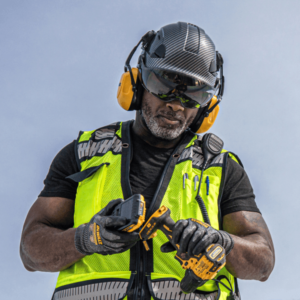 MAN WEARING A DEWALT HI-VIS SAFETY VEST, GLOVES, SLATE COLORED TYPE II VENTED HELMET, SMOKE VISOR AND CAPMOUNT EARMUFFS ATTACHING BATTERY TO A DRILL